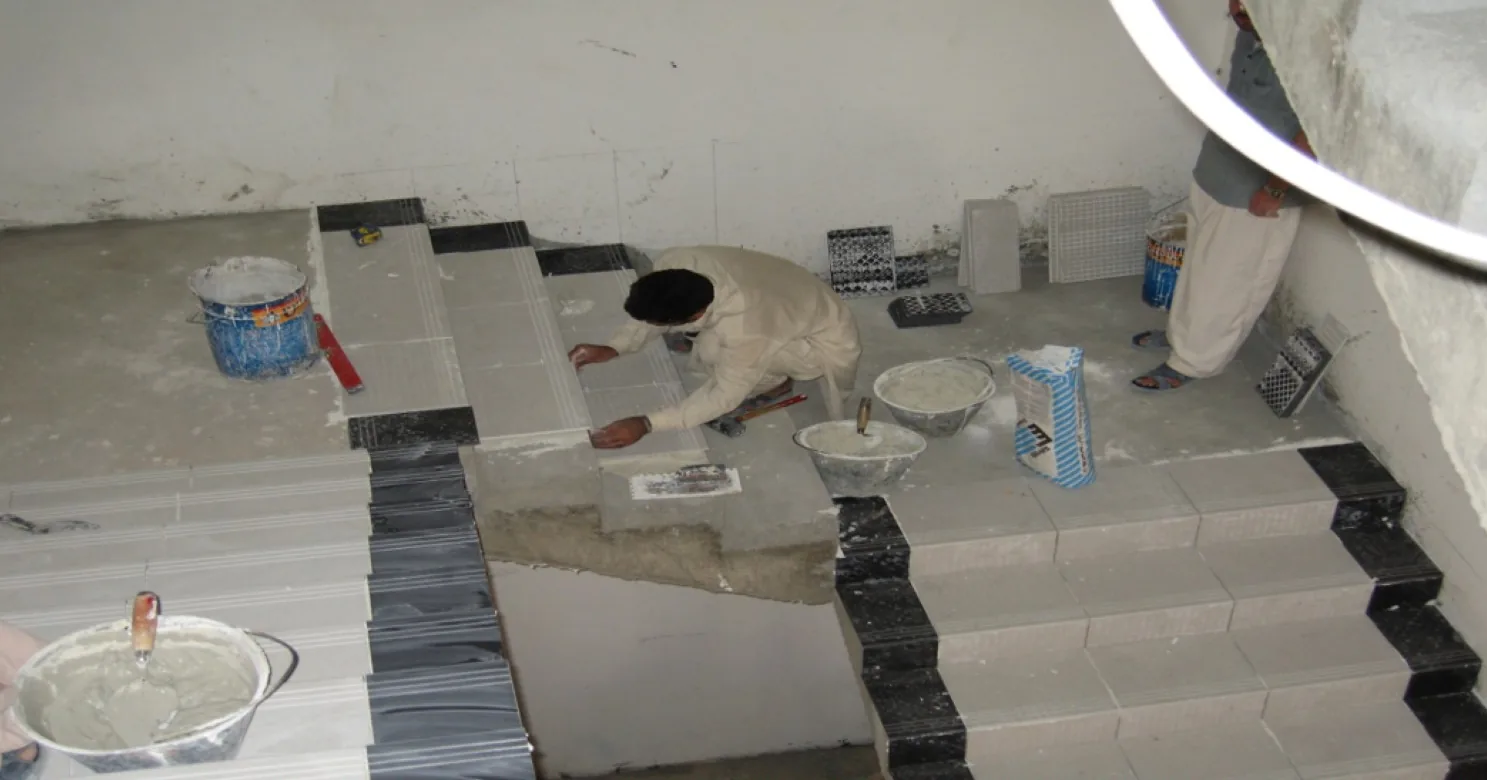 Worker tiling a concrete stair beside finished steps edged with black strips, with mortar buckets and an adhesive bag nearby