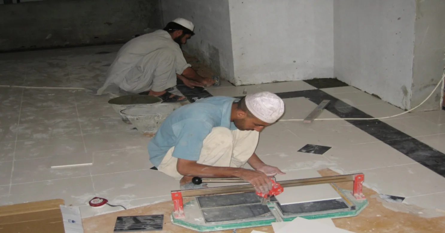 One installer cuts floor tile with a hand cutter while another sets tile beside a black inlay band in a bare concrete room