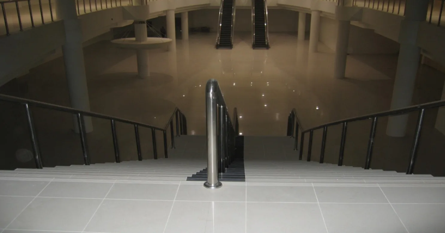 High view down a tiled staircase with black handrails and a steel post onto a dim, polished atrium floor with escalators beyond