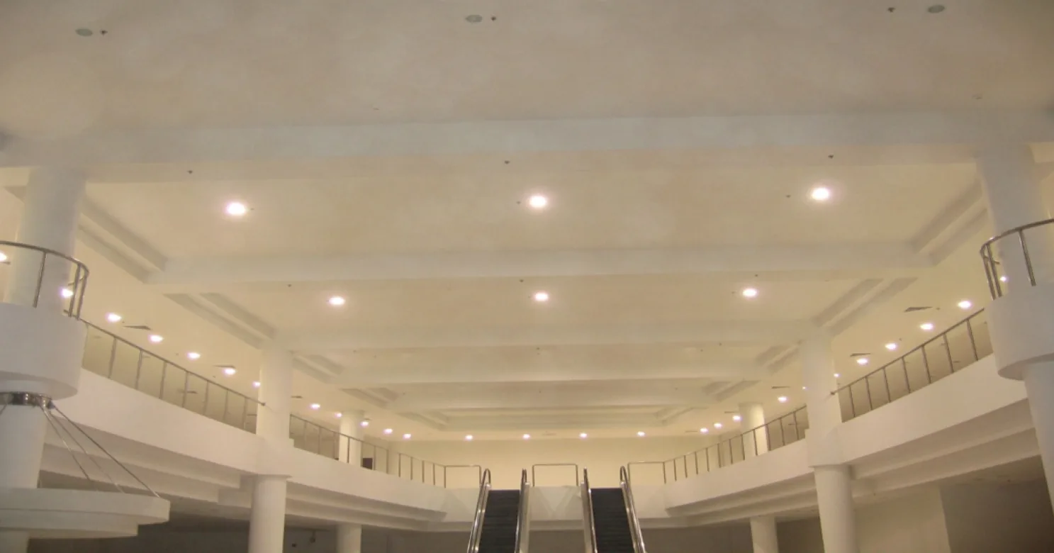 Looking up at a white beamed atrium ceiling with recessed downlights, above balustraded upper galleries, columns and a pair of escalators
