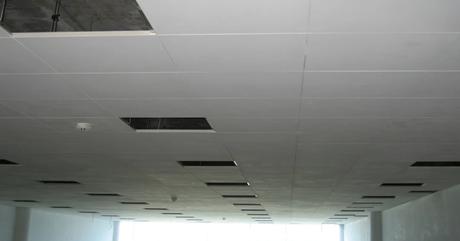 Suspended tile-grid ceiling with panels lifted out, linear air diffusers and a small sensor, running toward a bright window