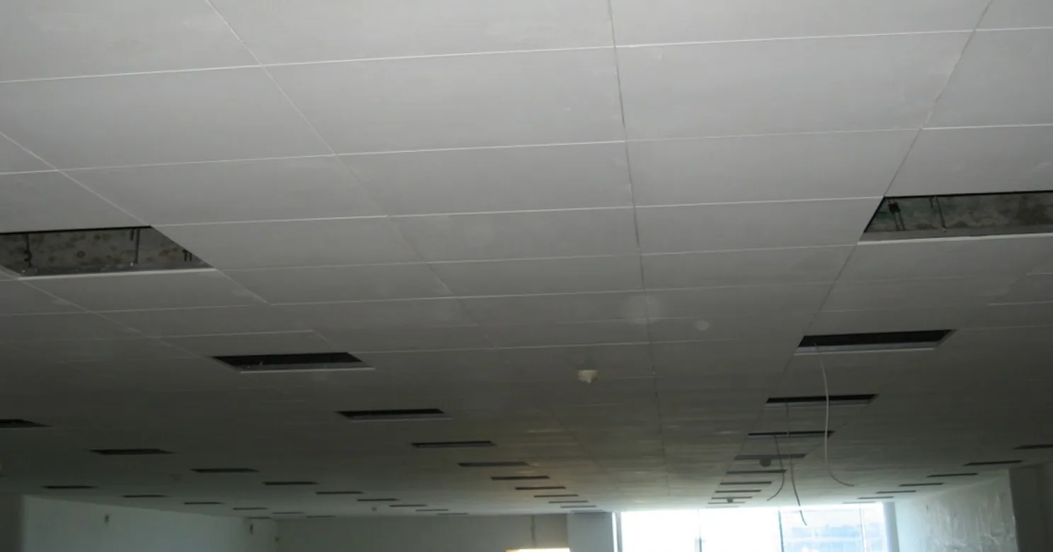 Suspended tile-grid ceiling with panels lifted out at both sides, loose cables hanging from an opening and rows of diffuser slots