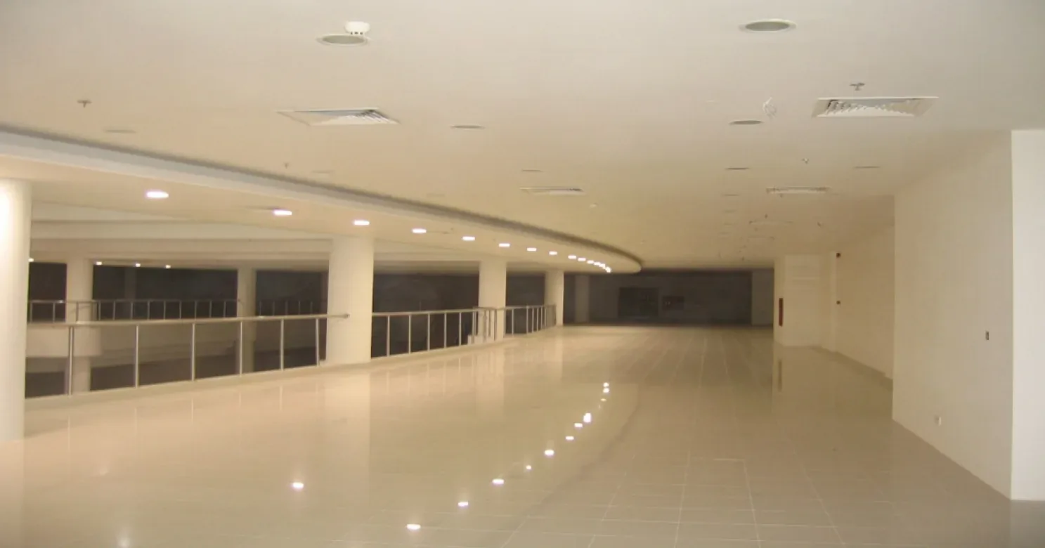 Empty curved upper-floor walkway of polished cream tile reflecting ceiling downlights, with a glass-and-steel balustrade and curved ceiling bulkhead