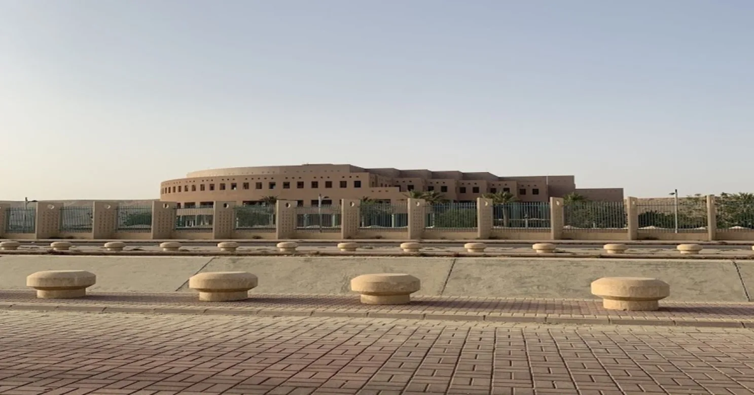 Distant round sand-coloured building behind a fence, seen across mushroom-shaped stone bollards and red interlock paving under a hazy sky.