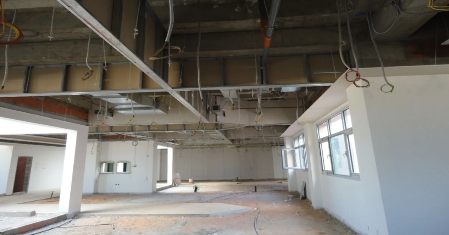 Unfinished open-plan floor with rough sandy ground, white walls and windows, and an exposed ceiling of ducts, steel framing and hanging cables.
