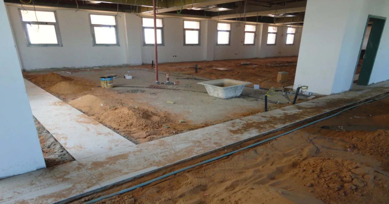 Large hall with the floor being prepared: sand mounds, pale screed strips, a vertical pipe riser, blue conduit and a white tub.