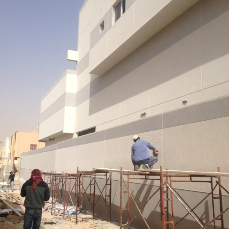 Wide view of a two-tone textured render elevation, scaffolding along its base and a worker crouching on the plank, work in progress.