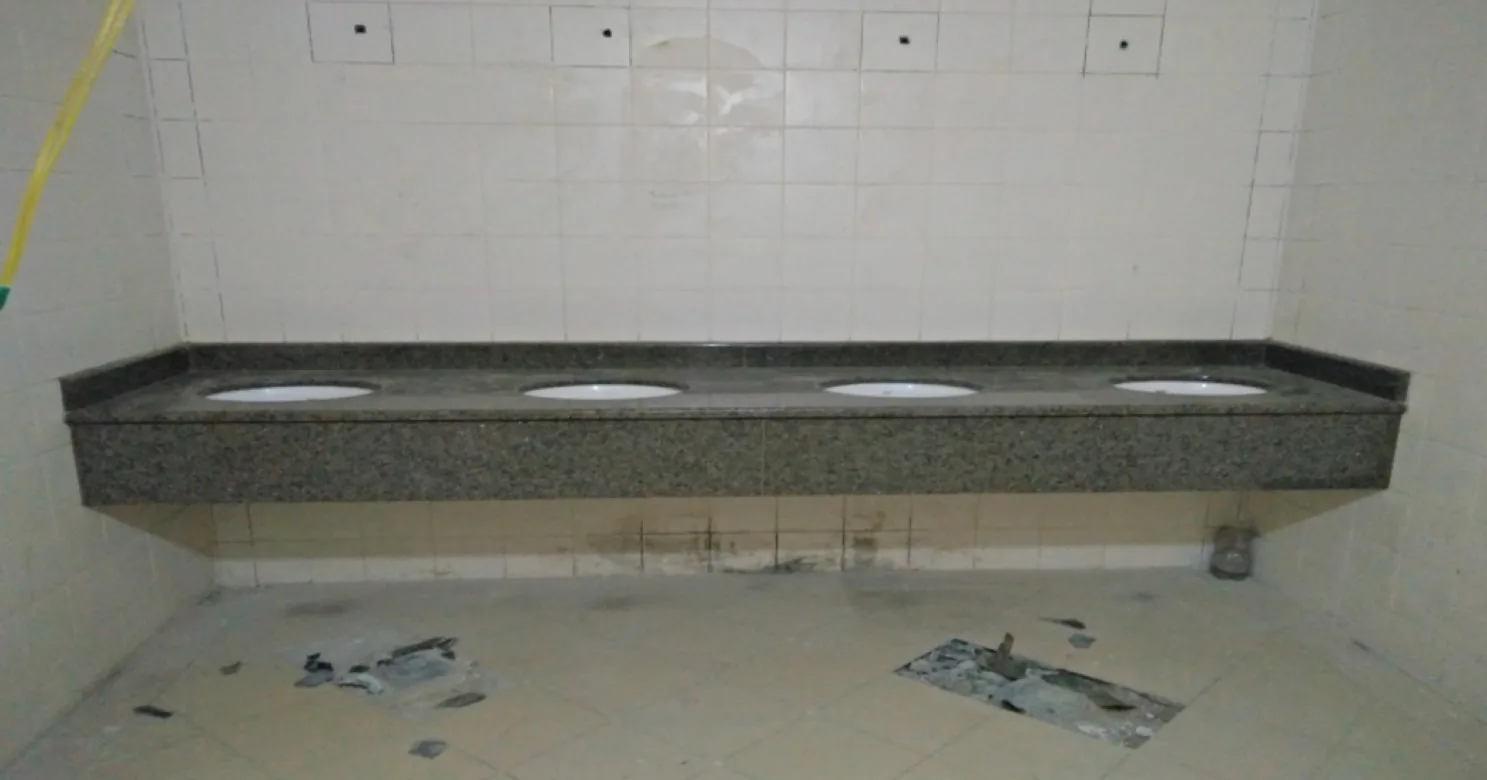 Four-basin granite vanity against a white-tiled wall, without taps fitted, with open floor cut-outs and debris in the tiling.