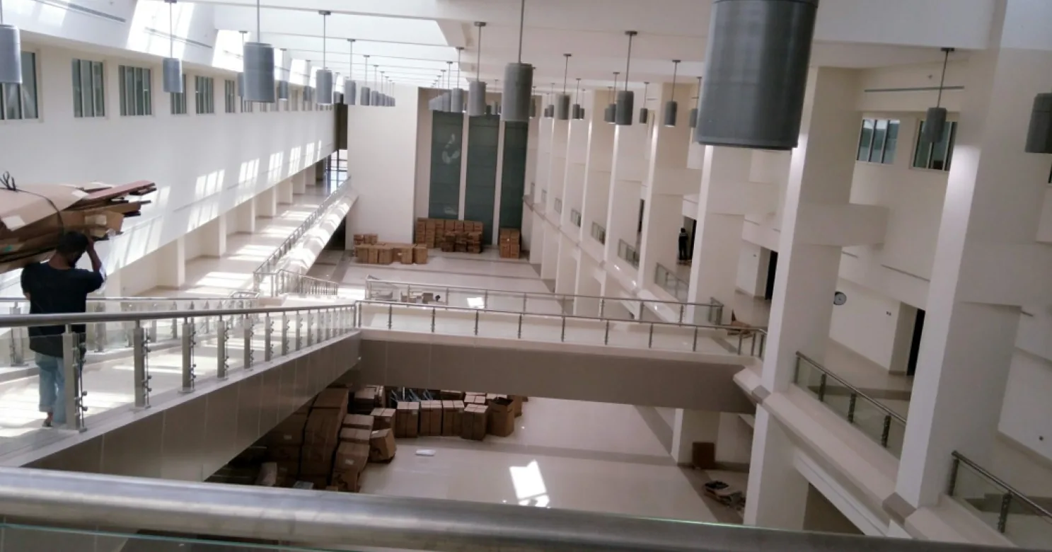 Atrium viewed from a mezzanine rail, with pendant cylinder lights, glass balustrades and stacked cartons below; a man carries a wrapped bundle.