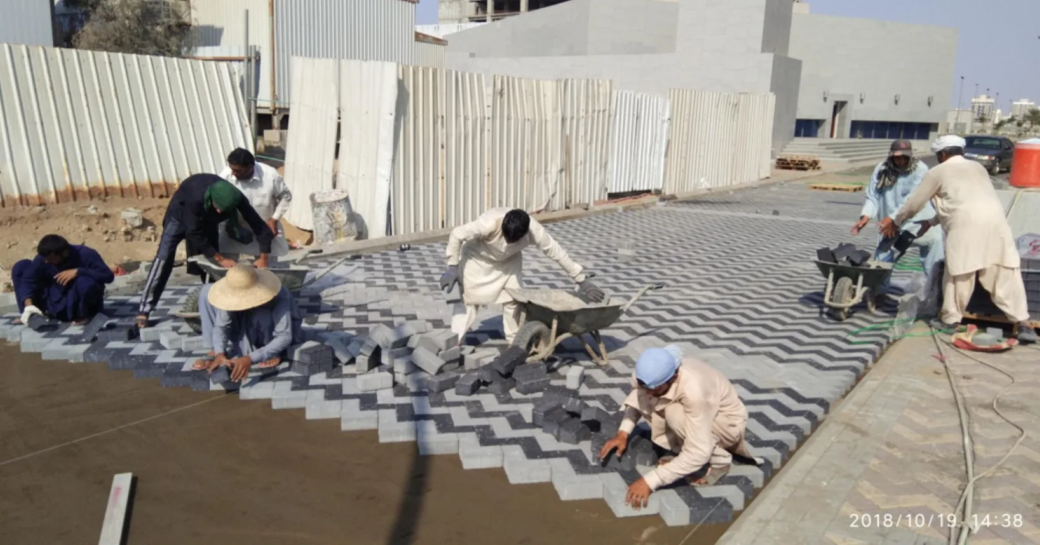 Crew laying grey and black chevron interlock pavers on a sand bed beside a corrugated site fence, with wheelbarrows and stacked pavers.