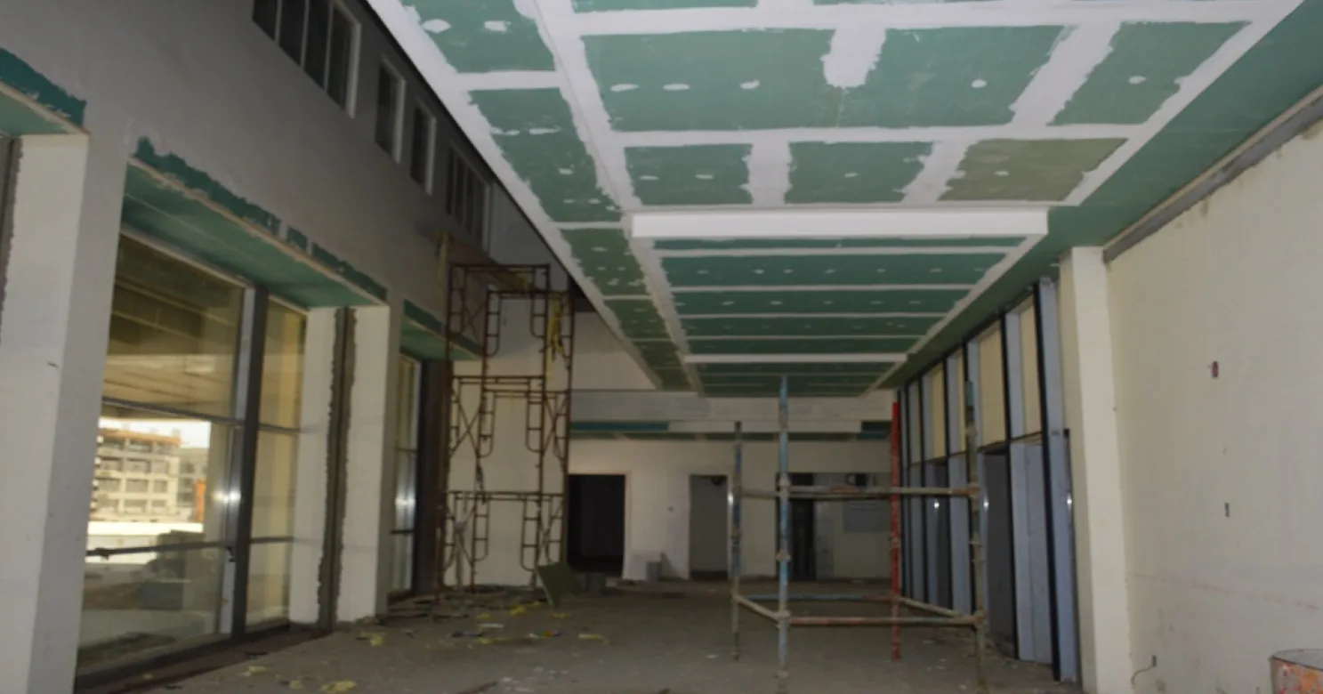 Corridor ceiling of green plasterboard being fixed and taped, with scaffolding, bulkheads, glazing frames and debris on the floor.