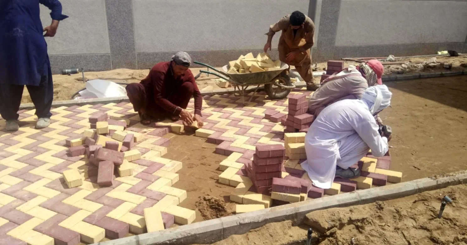 Workers laying yellow and maroon herringbone pavers on a sand bed along a kerb, with a grey stone-clad boundary wall behind.