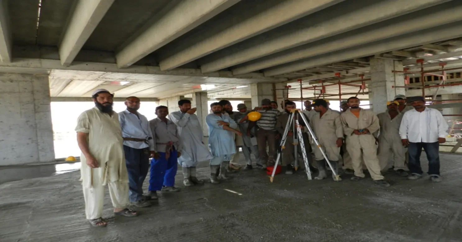 A large group of workers standing together on a concrete deck beneath ribbed concrete beams, with a surveying instrument on a tripod.