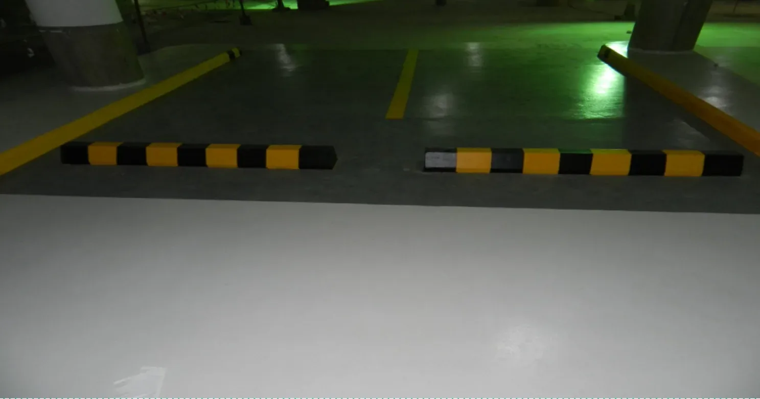 Wider view of a basement parking bay with a pale floor, yellow lines, wheel stops and concrete columns under dim green-tinted light.