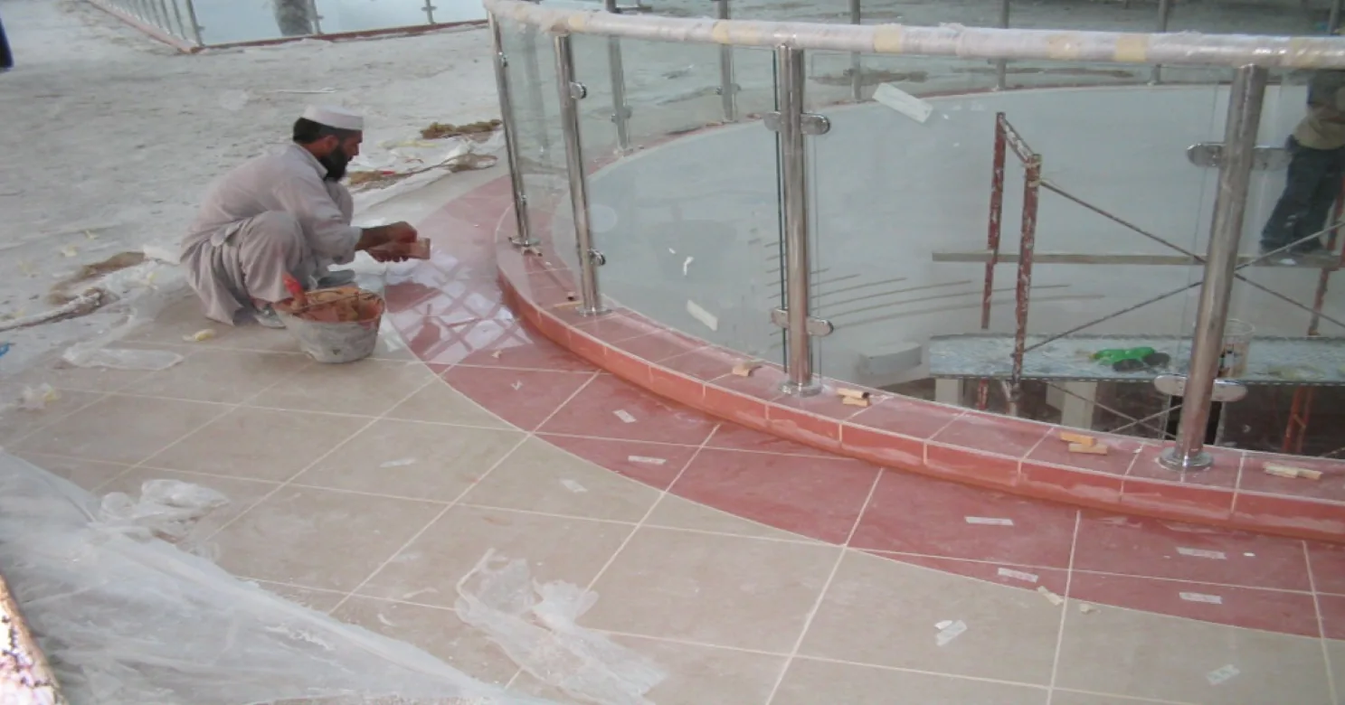 An installer crouches beside a bucket, fixing floor tile next to a curved terracotta band, with a glass-and-steel balustrade and plastic sheeting