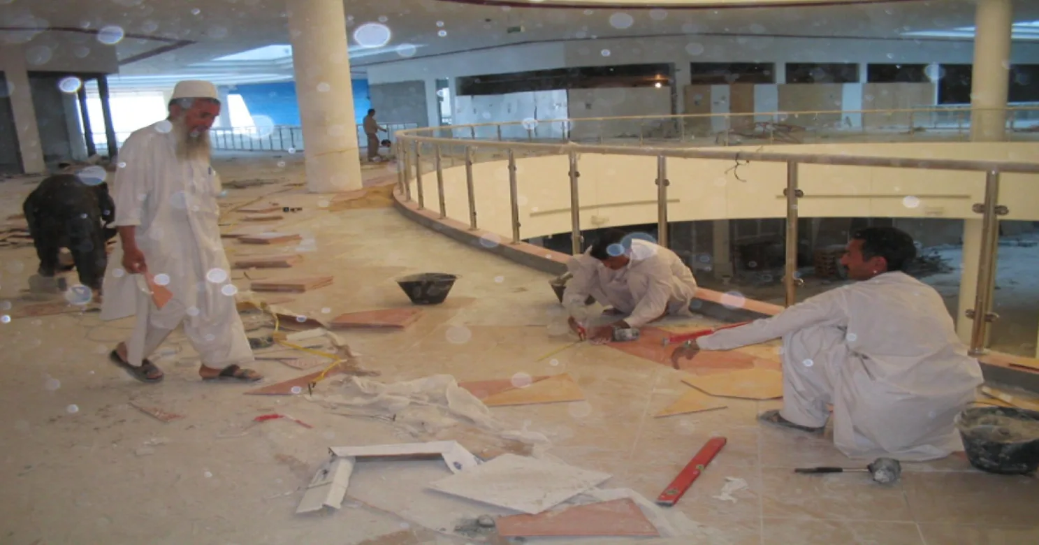 Five workers on a mezzanine, three crouched over floor tile beside a curved balustrade, offcuts and tools scattered around