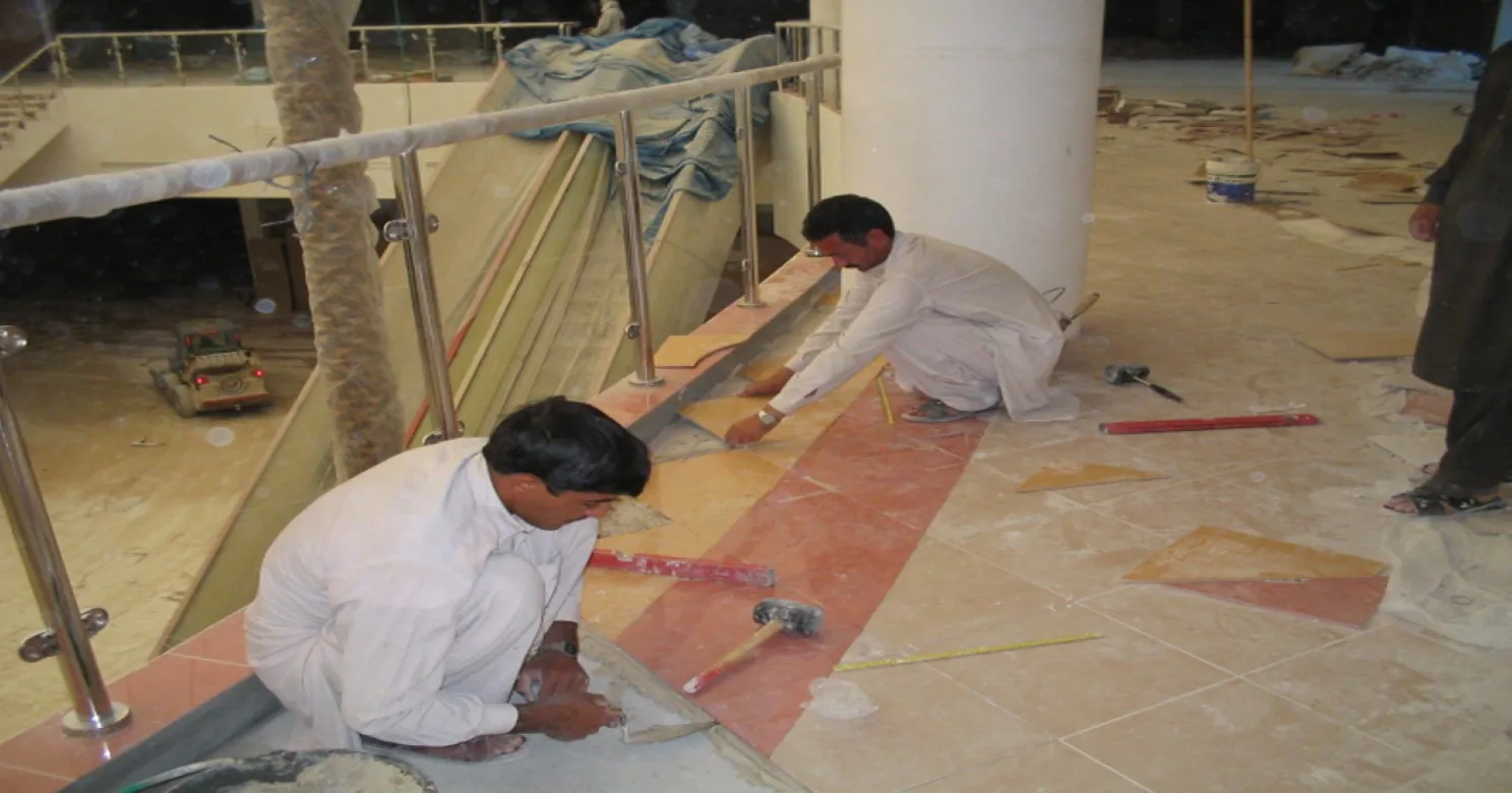 Two installers set ochre and terracotta floor pieces along a curved band, with a spirit level, mallet and tape measure nearby