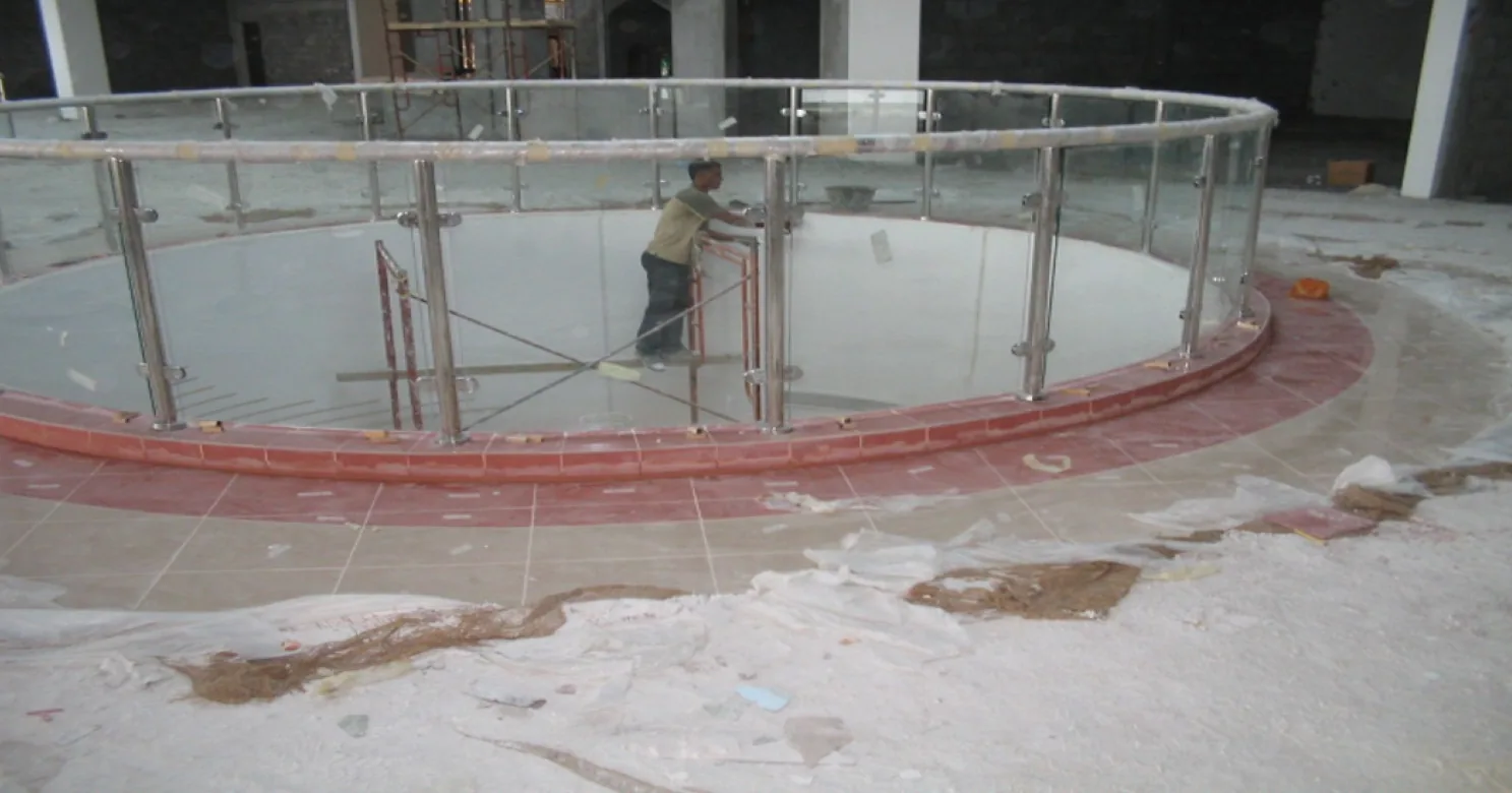 Circular glass balustrade around a void, a worker on scaffold inside, terracotta tile band at its foot, debris in front
