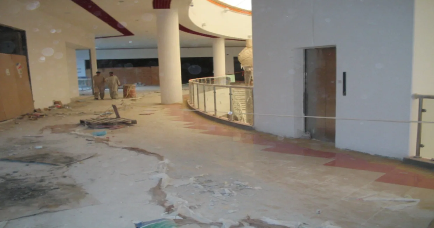 Partly tiled mezzanine walkway with a terracotta zig-zag band along a curved balustrade, debris underfoot and two workers in the distance