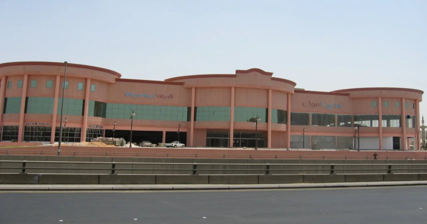 Pink two-storey mall facade with curved bays, glazed bands and Arabic and English signage, seen across an empty road