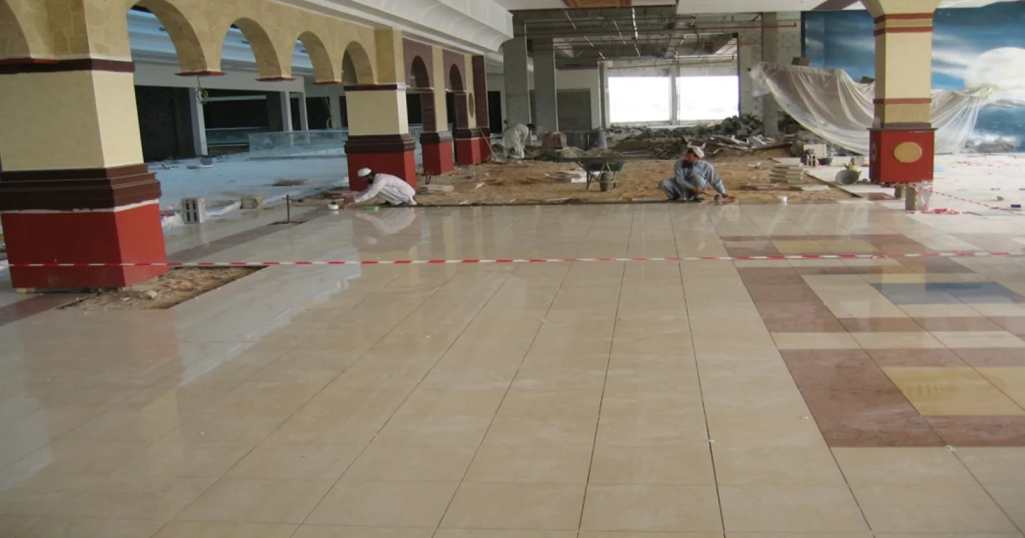 Polished cream floor tile along an arcade of painted arches and red-based columns, inlay squares at right, sand bed still bare beyond