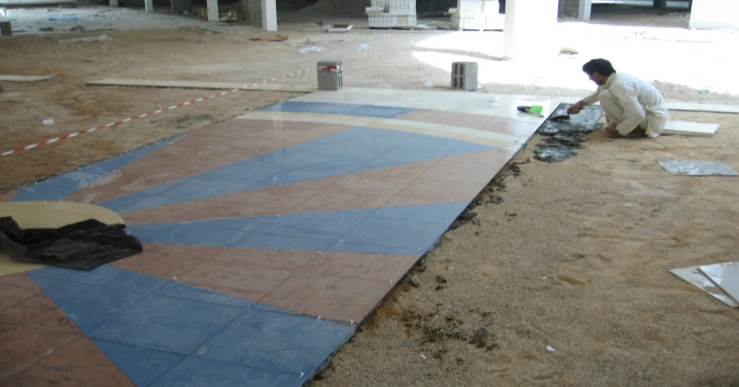 One installer kneels beside a fan-shaped medallion section of blue and terracotta tile laid on a bare sand bed