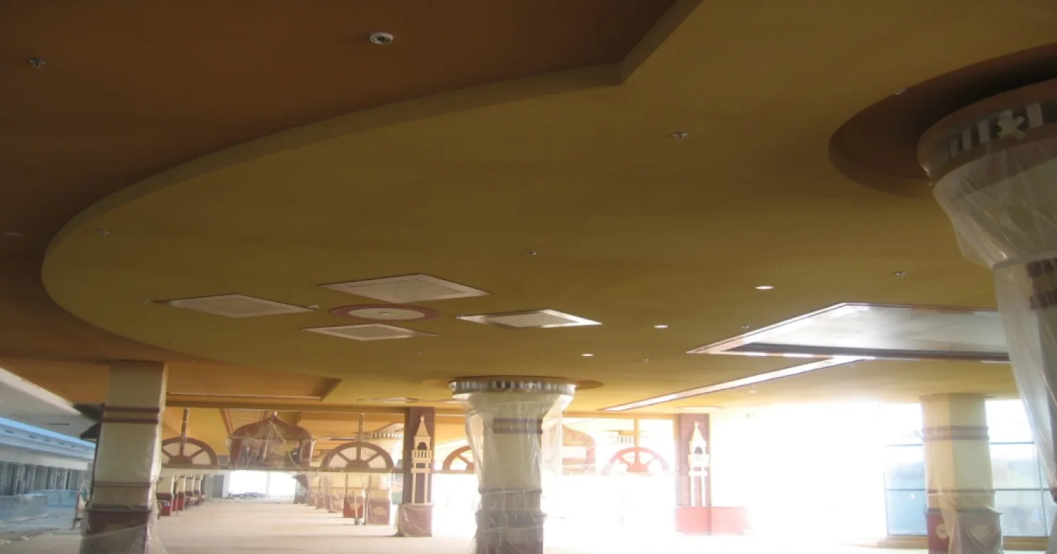 Curved yellow-painted suspended ceiling with small square air diffusers and recessed lights, above plastic-wrapped decorated columns, with strong daylight glare at right