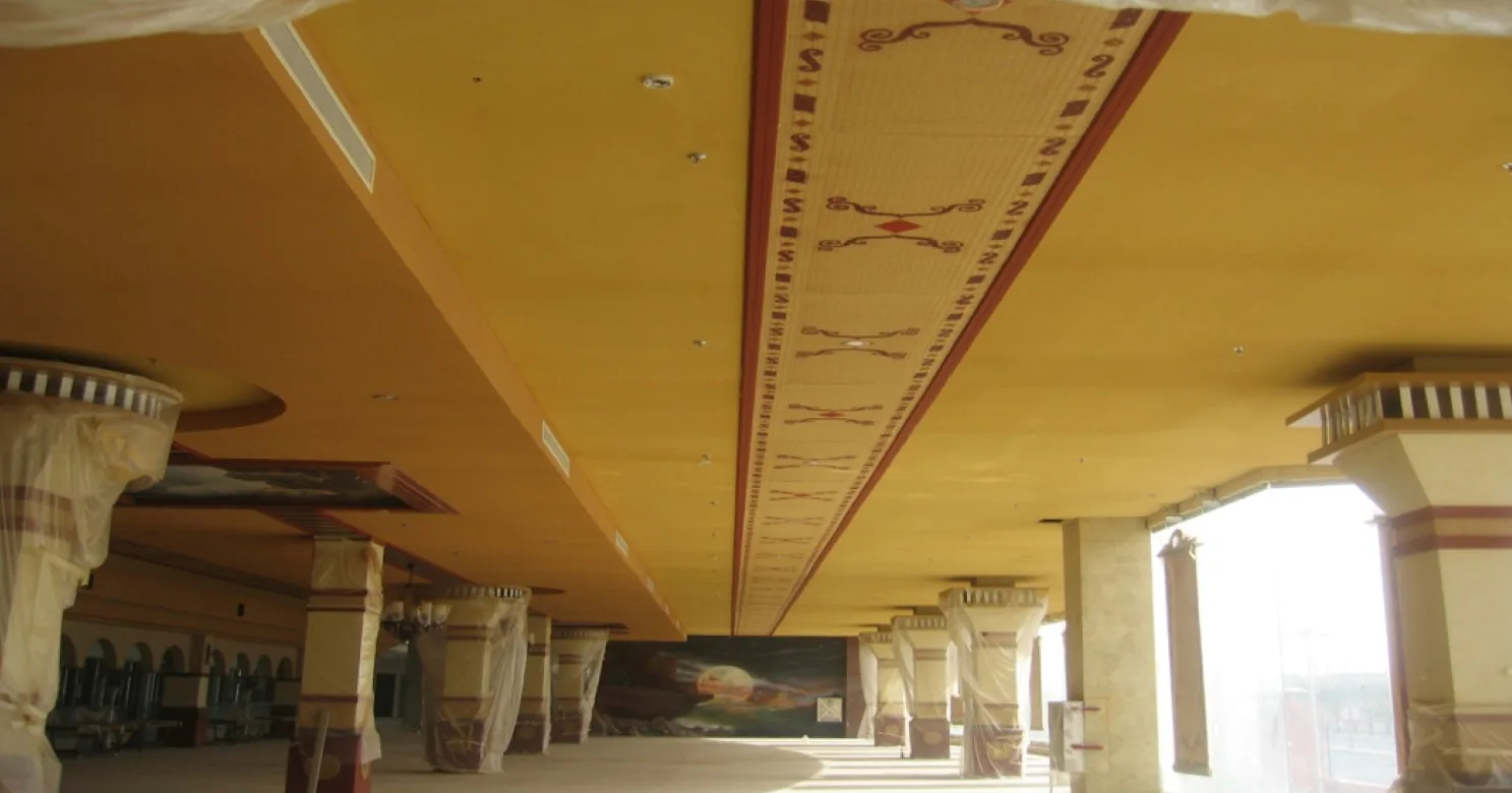 Yellow suspended ceiling with a central painted frieze of scrollwork and diamonds, over plastic-wrapped columns and a mural at the far end