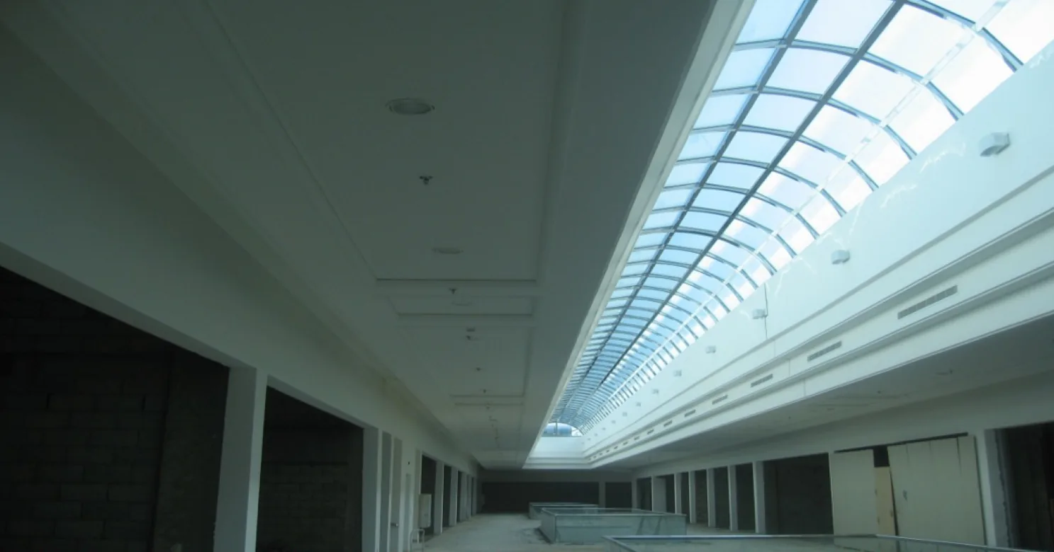 White suspended ceiling with recessed lights running beside a long arched glass skylight, above unfitted shop bays with bare block walls
