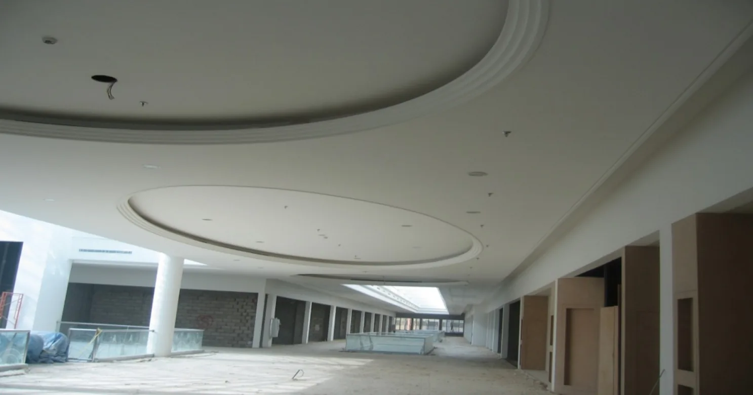 Two oval white ceiling recesses with moulded edges over a shell corridor, boarded shop openings at right, block walls at left