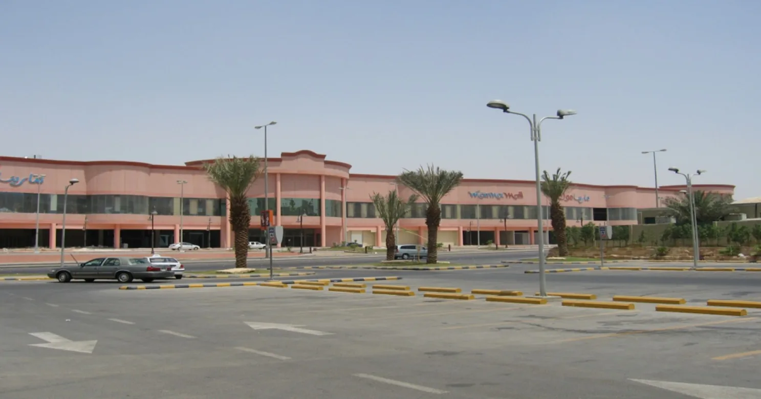 Long pink mall frontage beyond an empty car park, with palm trees, yellow-and-black kerbs, street lights and a car at left