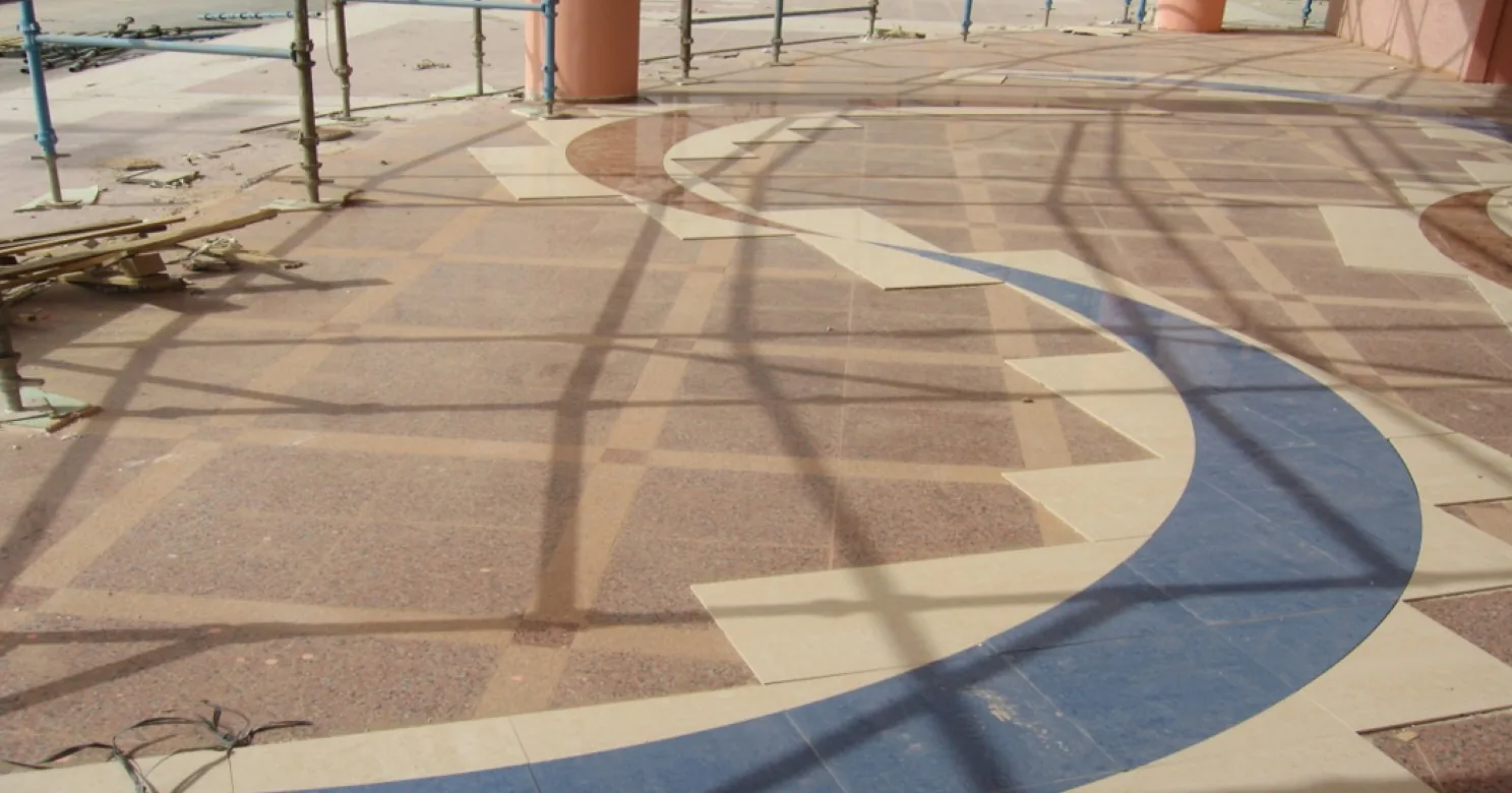 Blue curved band with stepped beige tile wedges set into a reddish speckled floor, scaffold shadows falling across the surface