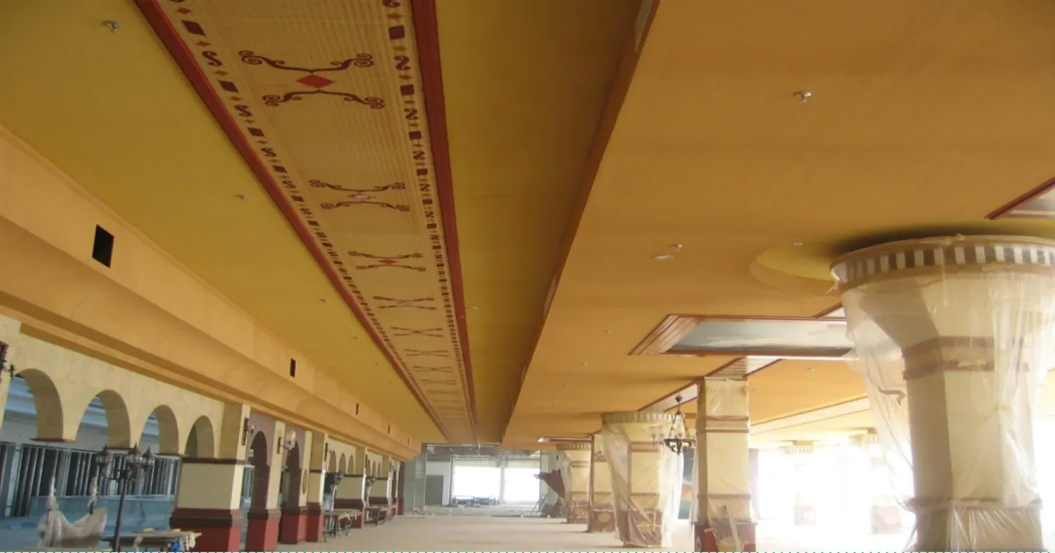 Corridor with a golden-yellow suspended ceiling and central painted frieze, arched bays at left and plastic-wrapped columns at right