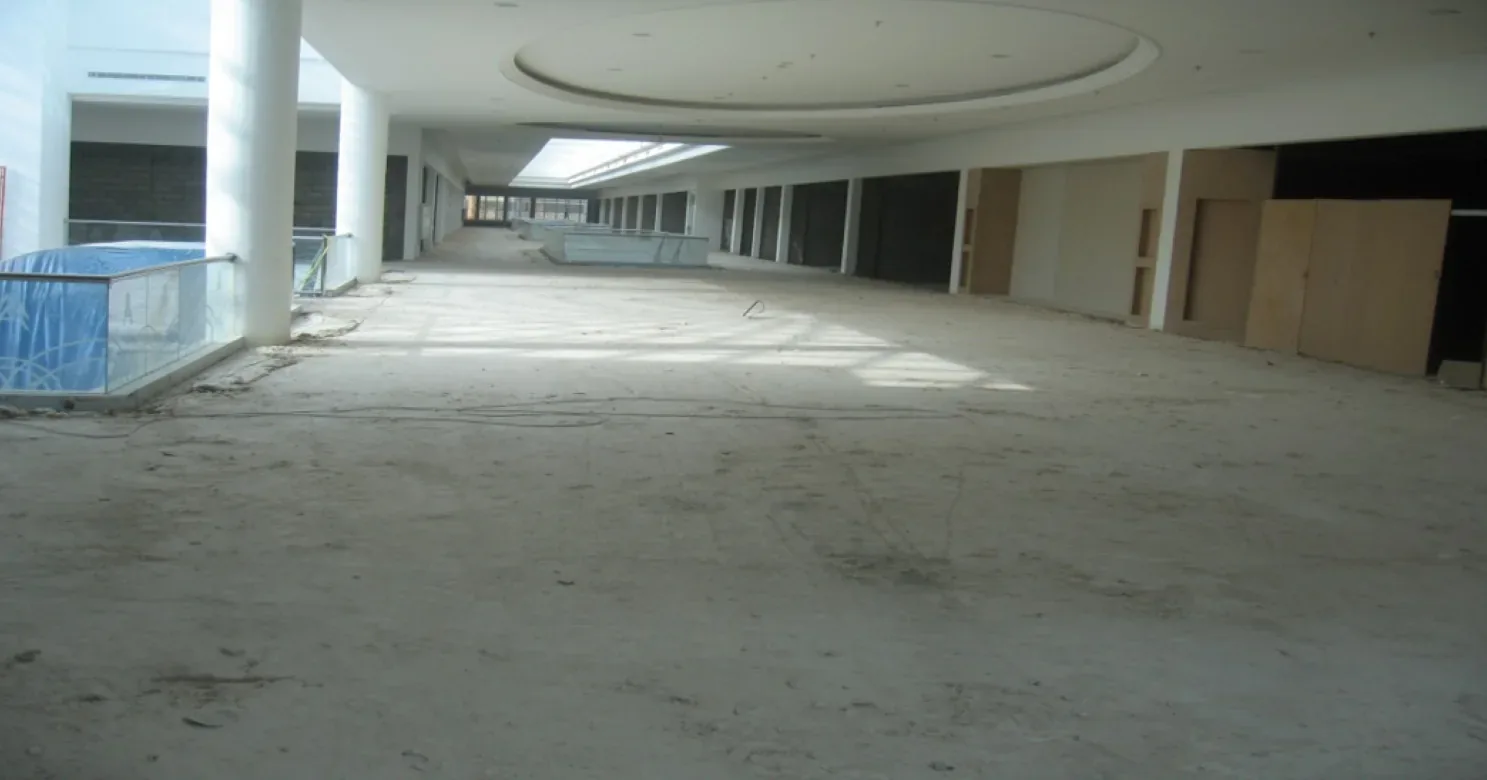Dusty bare concrete floor along a shell corridor with an oval ceiling, blue-wrapped glass balustrades and boarded shop openings