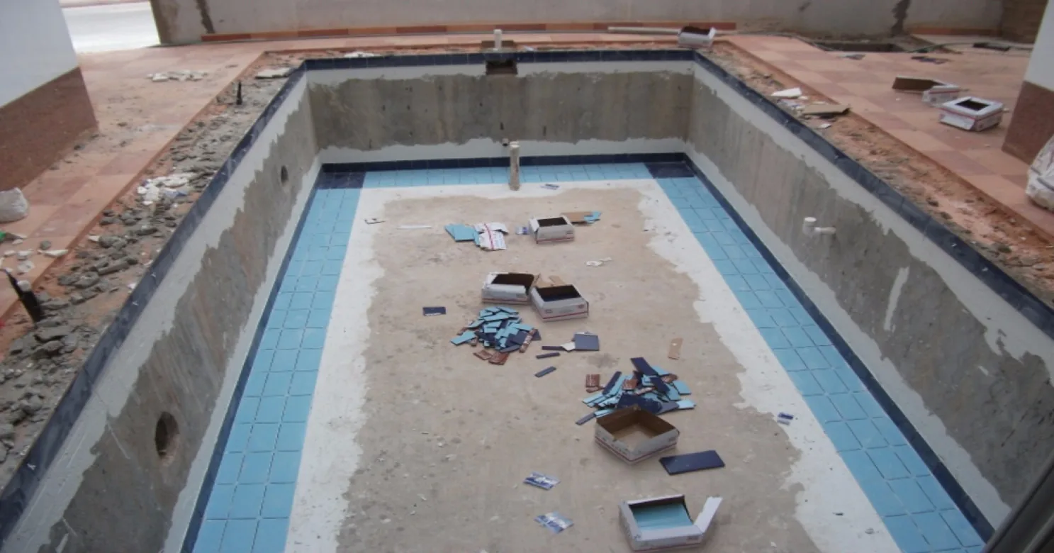 Pool shell mid-tiling: pale-blue tile bands along the floor edges, dark-blue border, bare centre, tile boxes and offcuts scattered.