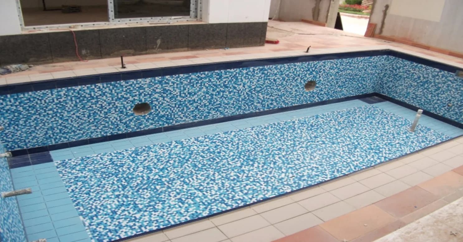 Empty pool fully lined in blue-and-white mosaic with dark-blue border tiles, cream and terracotta paving around it still being completed.