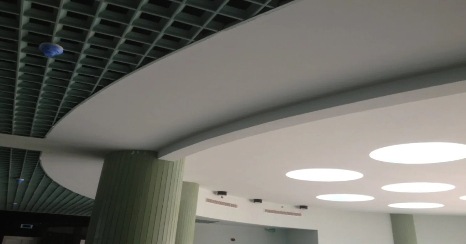 Curved suspended gypsum ceiling with bright round openings beside a green open-cell metal ceiling and green fluted columns, interior finished.