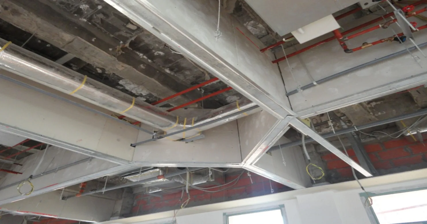 Open ceiling with steel-stud framing and gypsum board boxing around insulated ducts and red pipes, brick wall behind, work in progress.