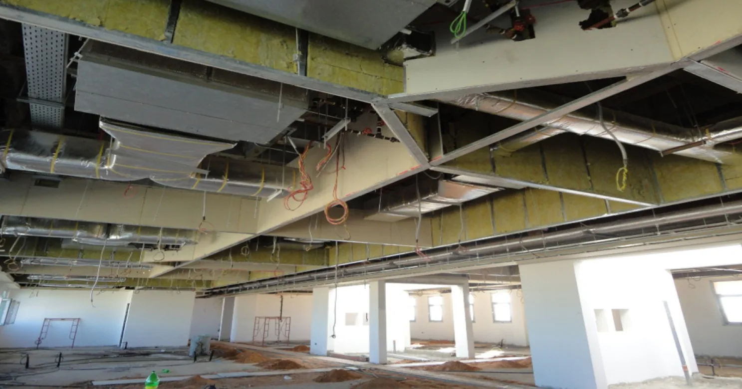 Wide open-ceiling floor with insulated ducts, perforated cable tray and gypsum boxing overhead, sand mounds and scaffold frames below, work in progress.
