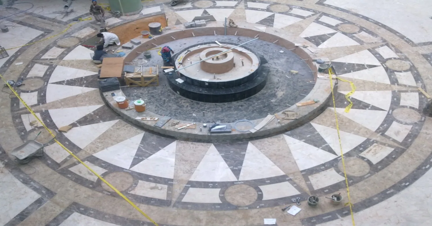 Overhead view of radiating marble floor pattern around a dark tiered fountain basin still under construction, with workers, tools and caution tape.