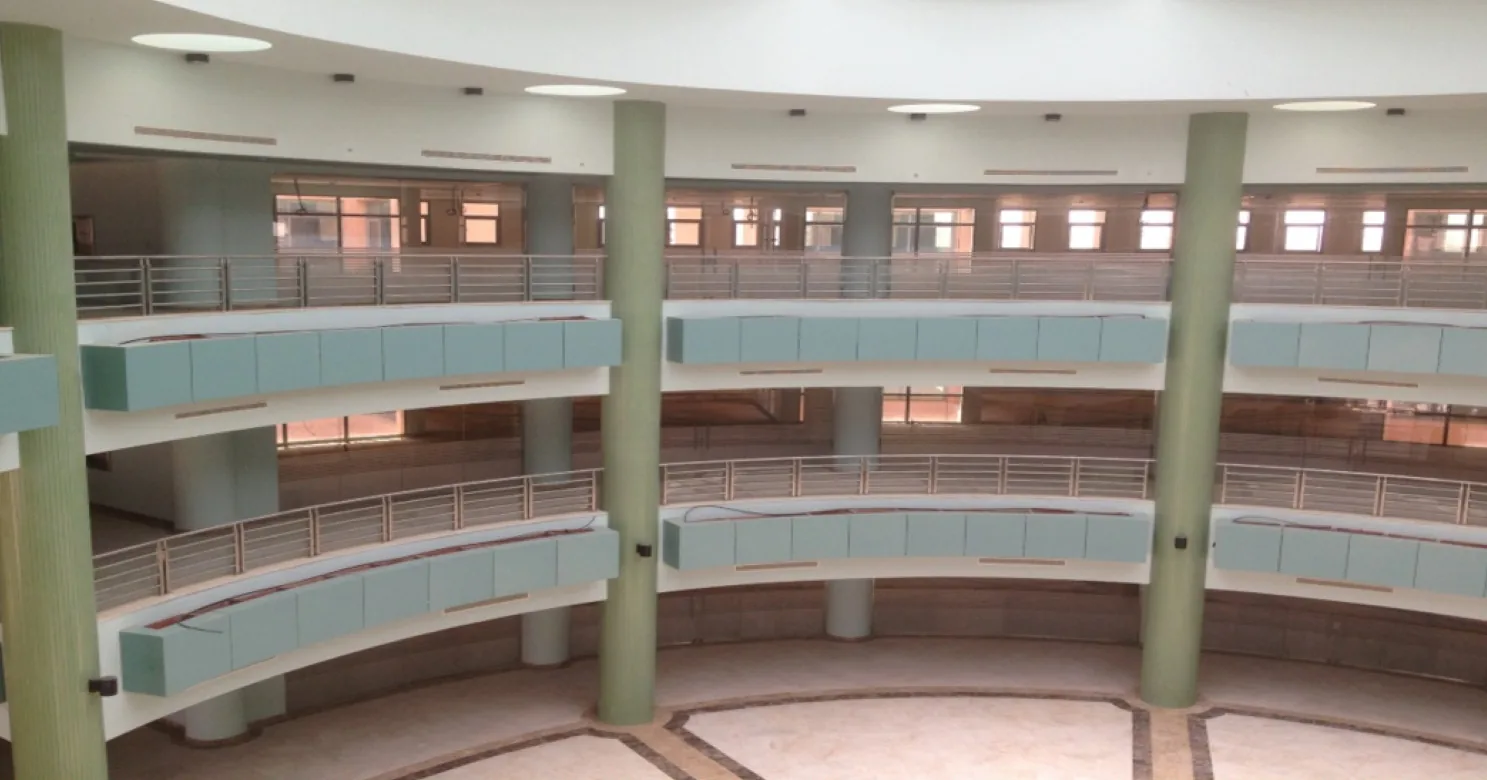 Wide view of a curved multi-storey atrium with green columns, steel-bar railings, pale-green balcony panels and a patterned marble floor.
