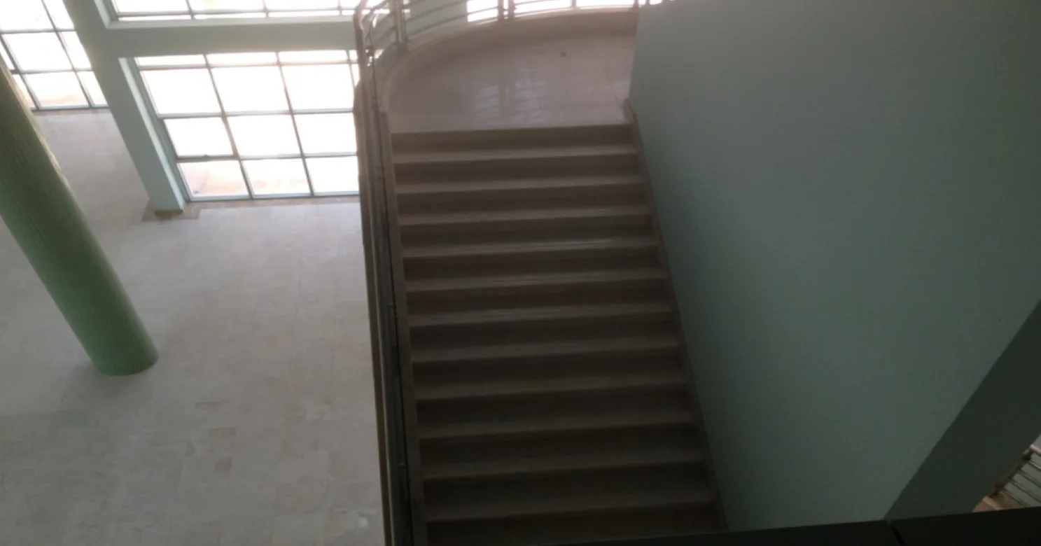 High view down a dim, backlit stair flight beside a green column and glazed wall, with a curved railing at the landing.