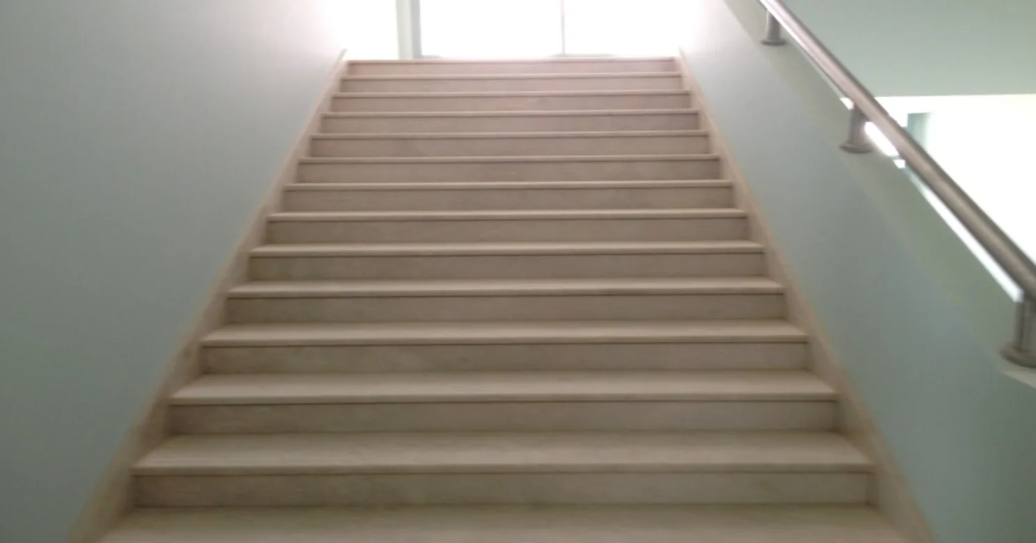 Straight stair flight with pale stone treads between light-teal walls, a steel handrail at right and bright light at the top.