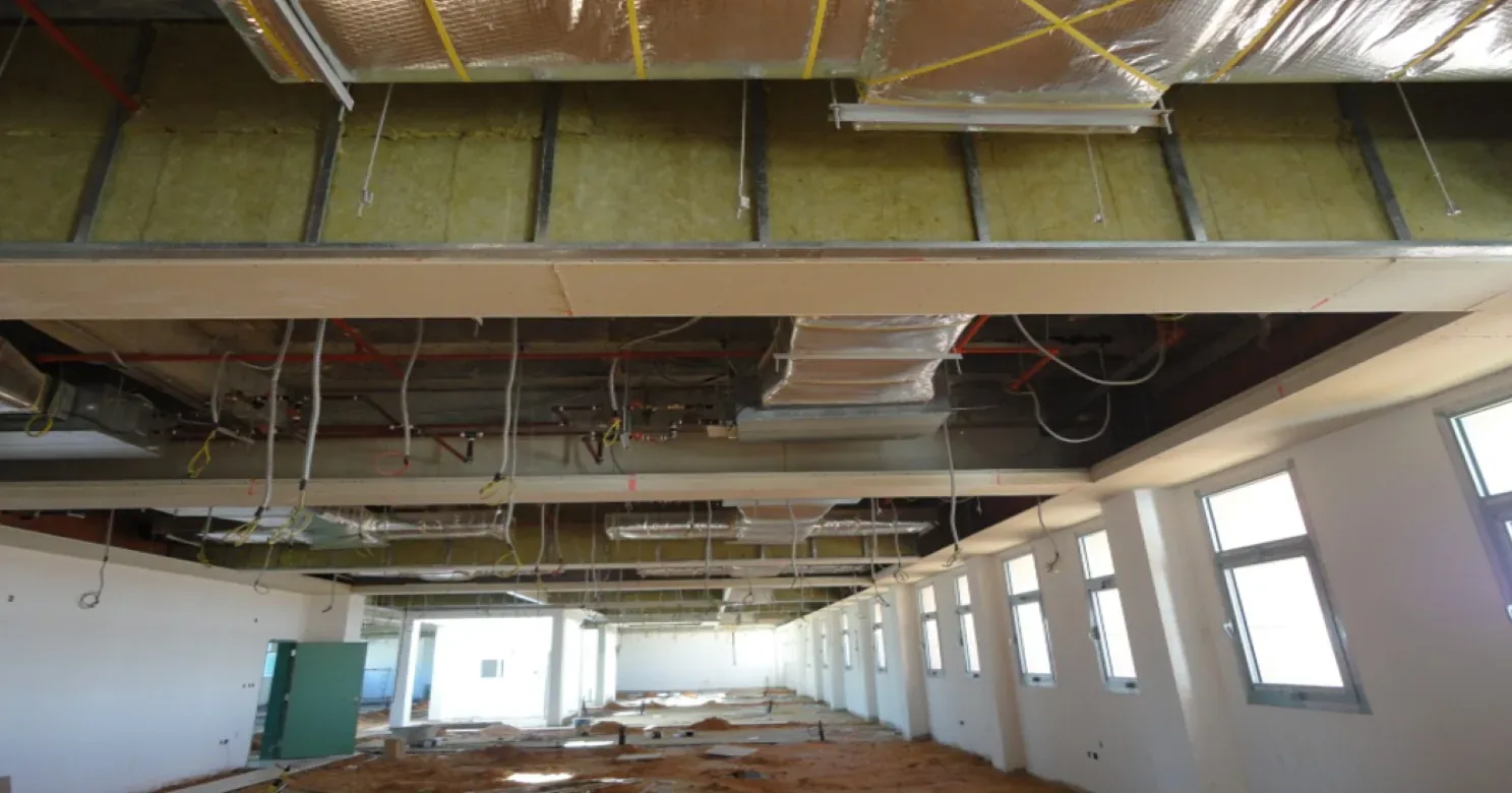 Open-ceiling hall lined with yellow mineral-wool insulation in steel framing, foil-wrapped ducts above, windows along one wall, sand-covered floor.