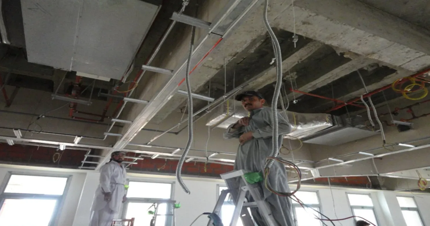 Two workers under an open ceiling, one on a stepladder handling flexible conduit and wiring beside cable tray, ducts and red pipes.