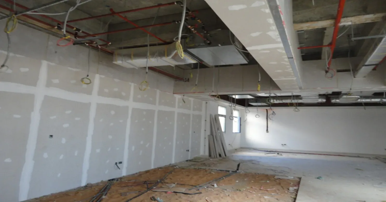 Gypsum board wall lining at the joint-filling stage beside an open ceiling with red pipes, wiring and a gypsum-boxed duct.
