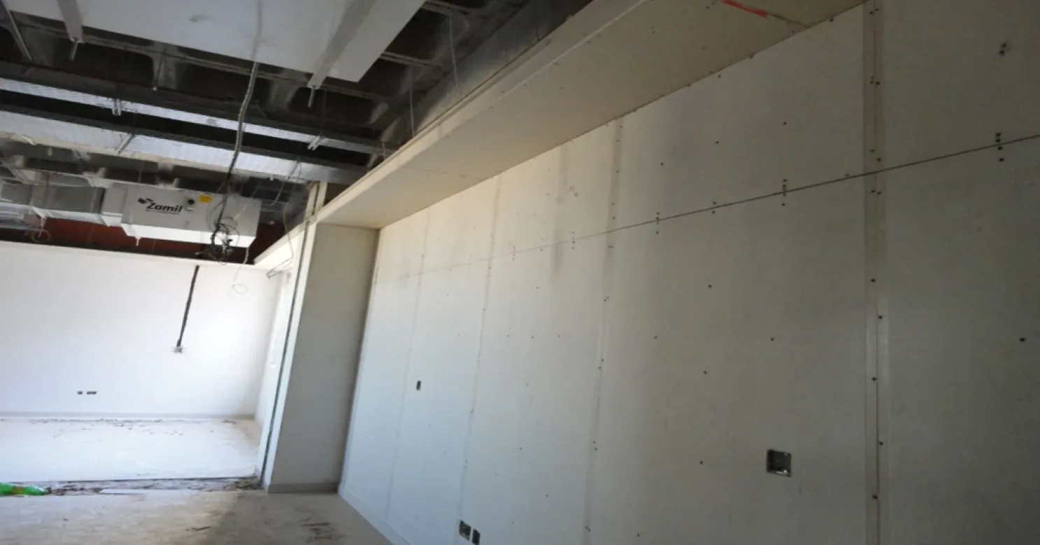 Unjointed gypsum board wall with visible screw heads and a boxed bulkhead above, an air-conditioning unit in the open ceiling behind.