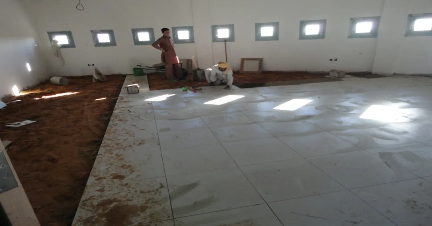 Large pale floor tiles being laid in a room with high square windows, one worker crouching, another standing by the bare base.