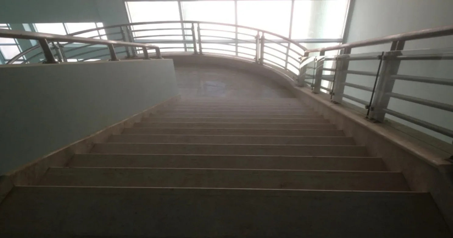 Stair flight rising to a curved landing with steel-and-glass railings under a glazed wall, seen in dim, hazy light.