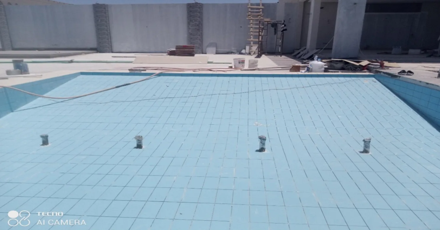 Wider view of the empty tiled pool with ladder, scaffold and building materials on the surrounding deck, boundary wall behind.